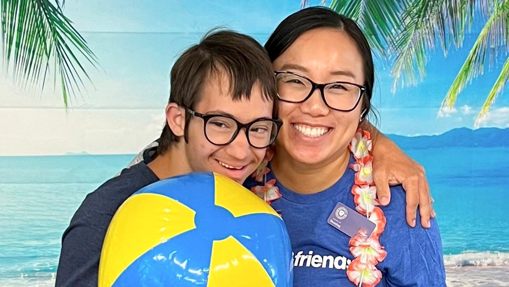 A boy and intern pose for a photo in front of a tropical beach backdrop. The boy holds a blue and yellow beach ball.