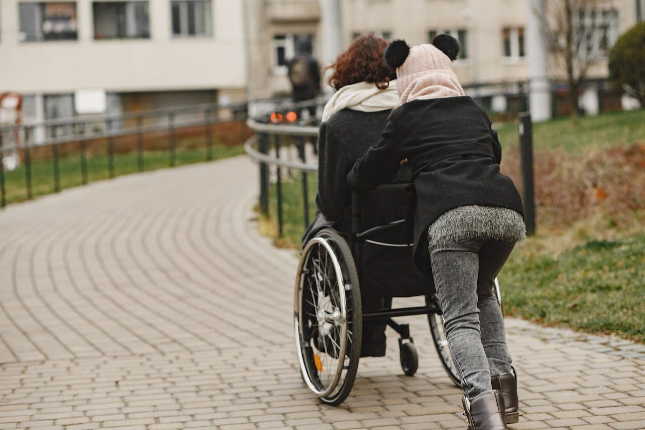 A person assisting someone else in a wheelchair while moving along a walkway.