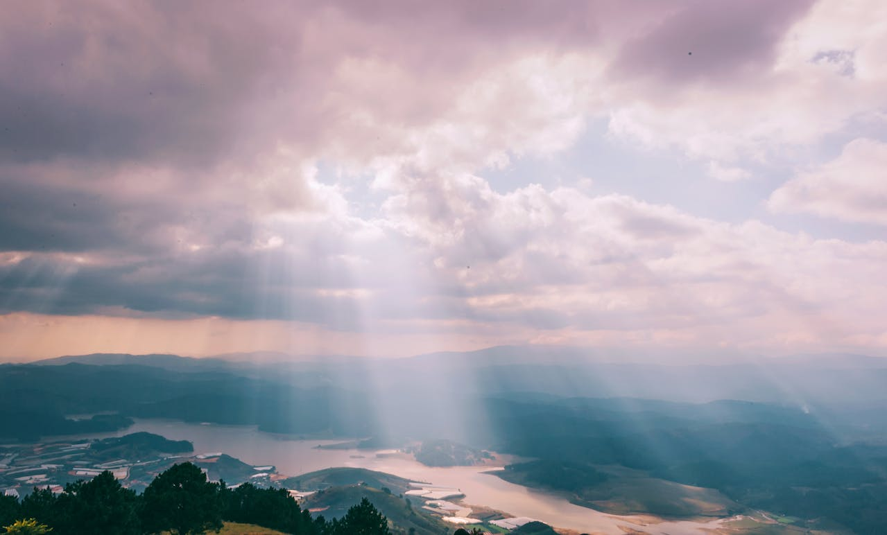 Sun rays breaking through clouds over a mountainous landscape with a river or lake below. 