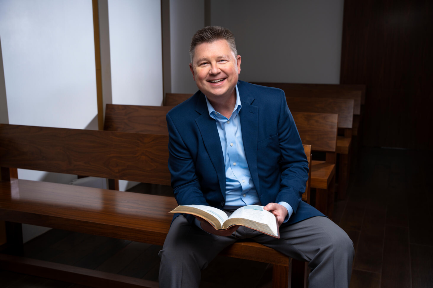 Steve Bundy is sitting on a bench inside a chapel, smiling for the camera with an open Bible.
