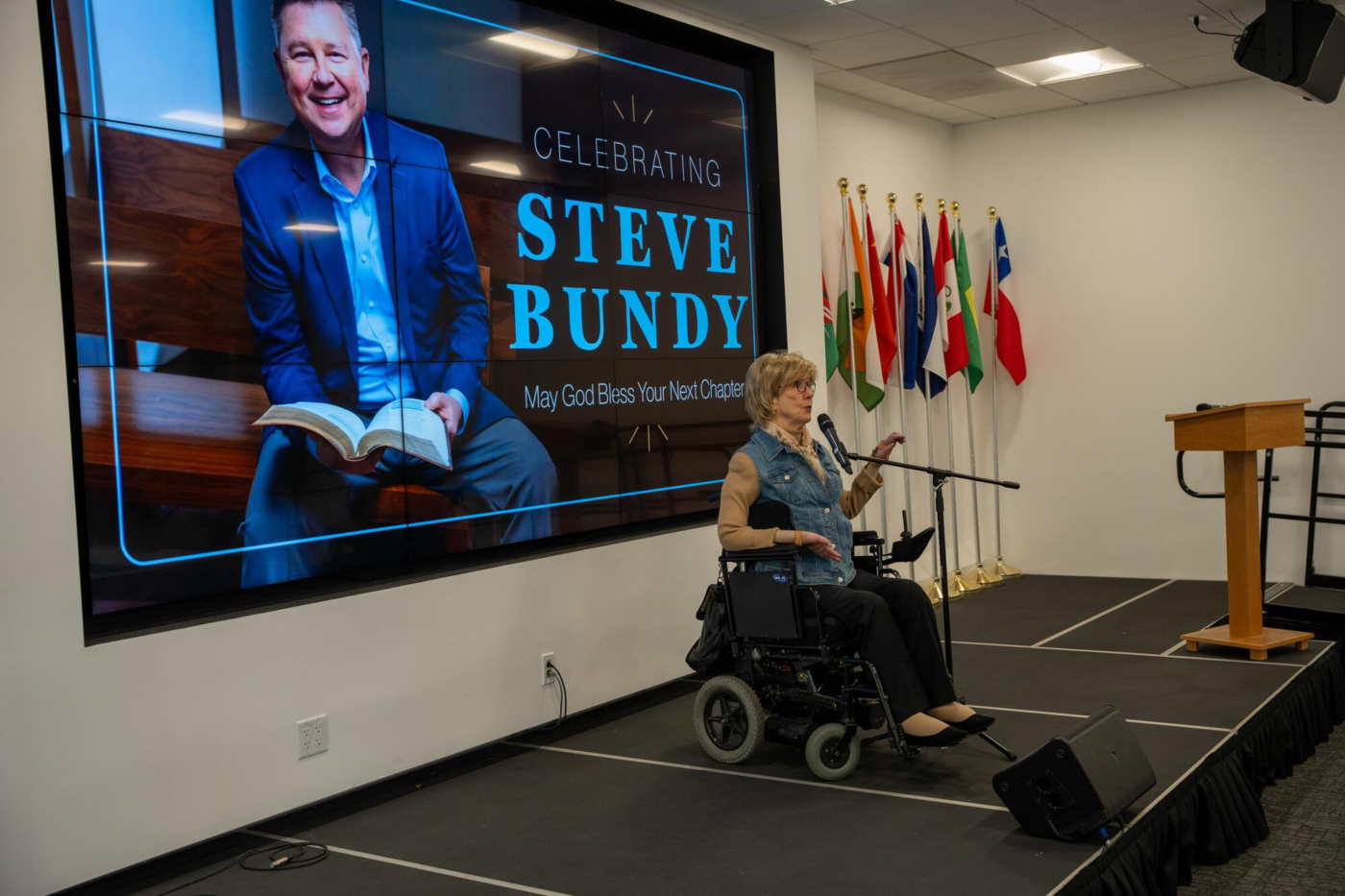 Joni Eareckson Tada speaking at a microphone on stage, with a screen behind her displaying a photo of Steve Bundy and the message "Celebrating Steve Bundy May God Bless Your Next Chapter."