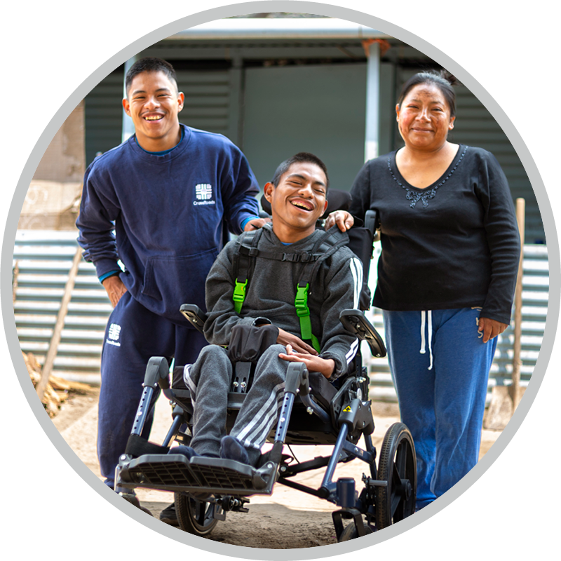Three people outdoors: a man in a wheelchair at center, with a man in blue sweats on his left and a woman in a dark top on his right, all smiling.