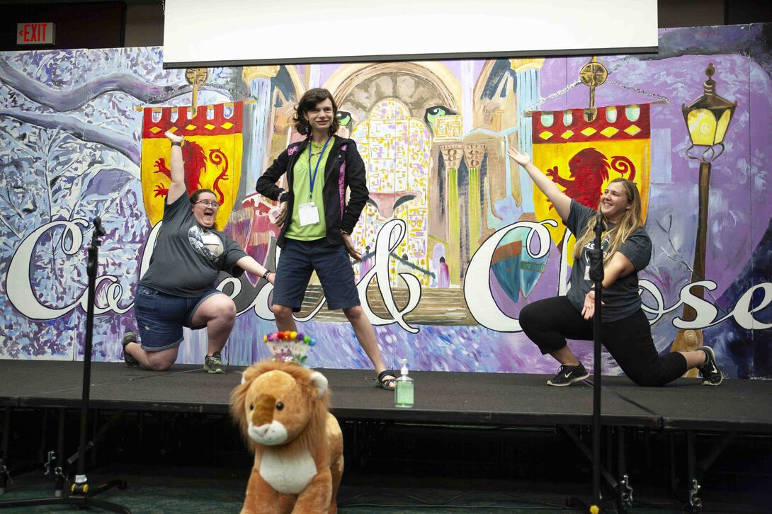 Katelyn poses on stage with two women kneeling beside her, their arms wide open as they present her.