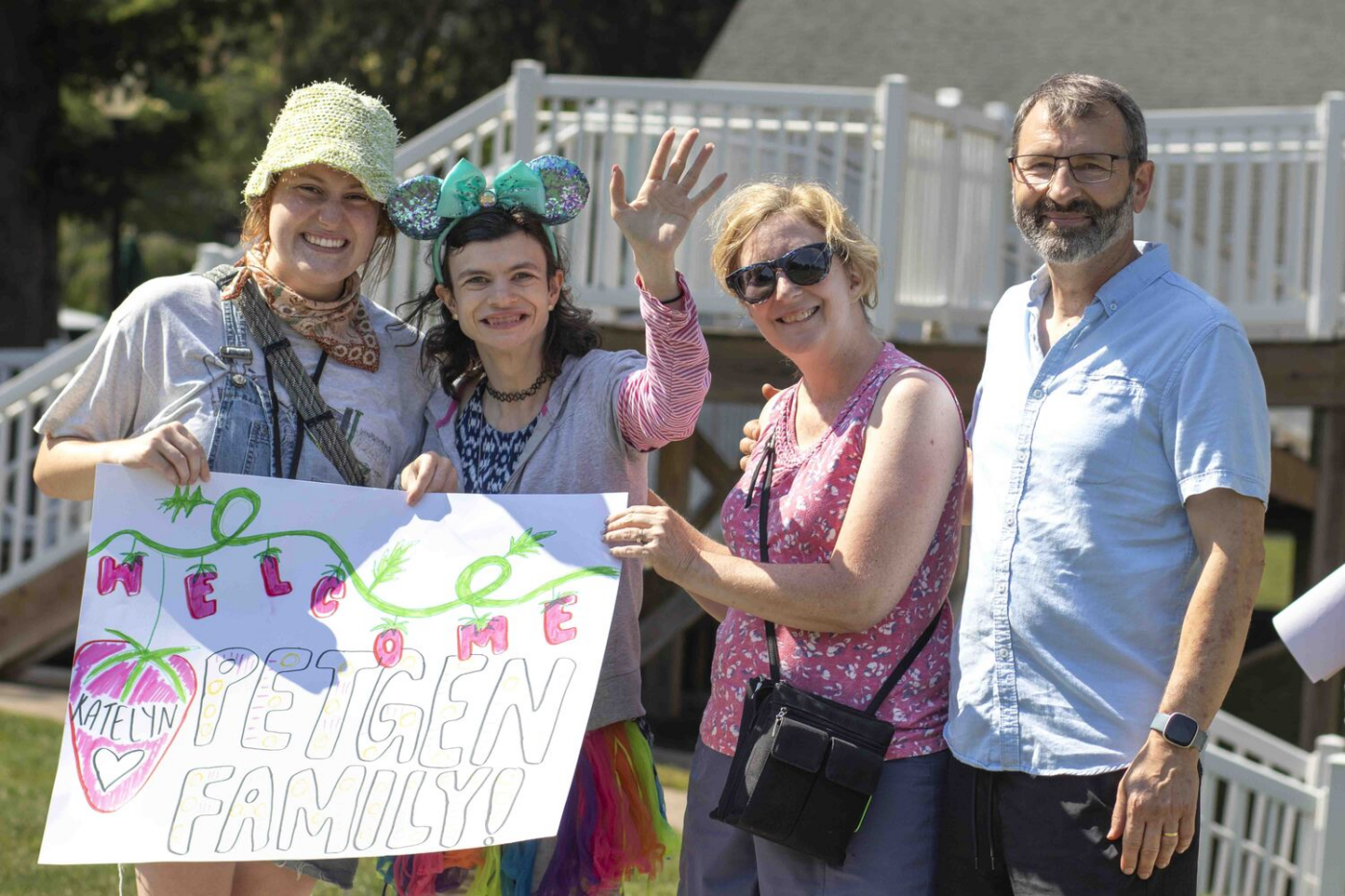 Four people stand outdoors, smiling while holding a colorful welcome sign that reads “WELCOME PETGEN FAMILY” with a heart-shaped graphic and the name Katelyn on it. Katelyn is waving.
