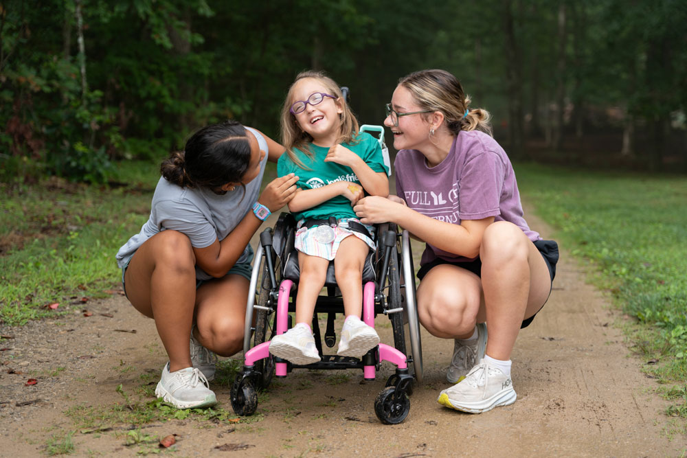 A smiling girl in a pink wheelchair sits between two women outdoors on a dirt path, all sharing a joyful moment.