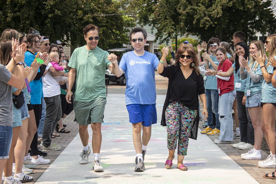 Yvonne, Ken, and their son Ryan walk hand-in-hand with raised arms as they are warmly welcomed by people clapping and smiling on both sides of the pathway.