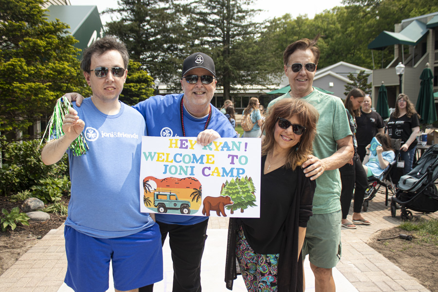 Ryan, Ken, and Yvonne stand with another man, smiling and posing for the camera as he holds a sign that reads "Hey Ryan! Welcome to Joni Camp."