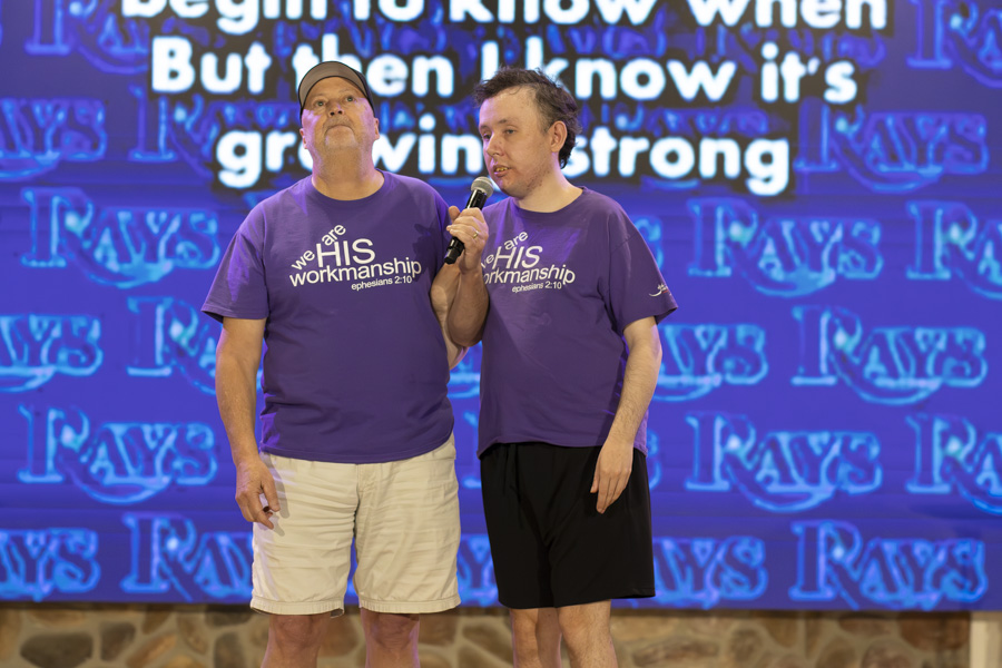 Ryan sings on a stage while his buddy holds the microphone; both are wearing shirts that say "We are His workmanship, Ephesians 2:10."