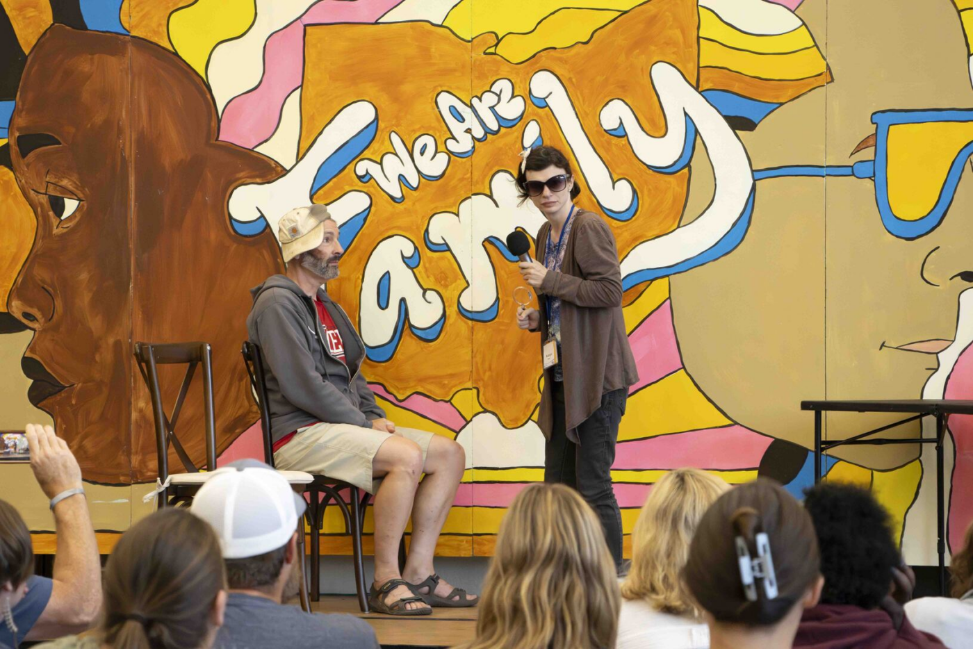 Katelyn stands on stage holding a microphone and looking toward the audience, with another person seated next to her on the stage.