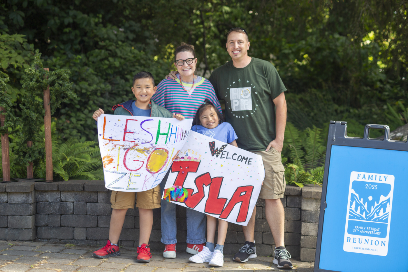 Molly, Justin, Isla, and Zeke at a family retreat, all posing and smiling for the camera. Justin is holding a sign that says "Leshhhh Go Zeke," while Isla is holding a sign that says "Welcome Isla."