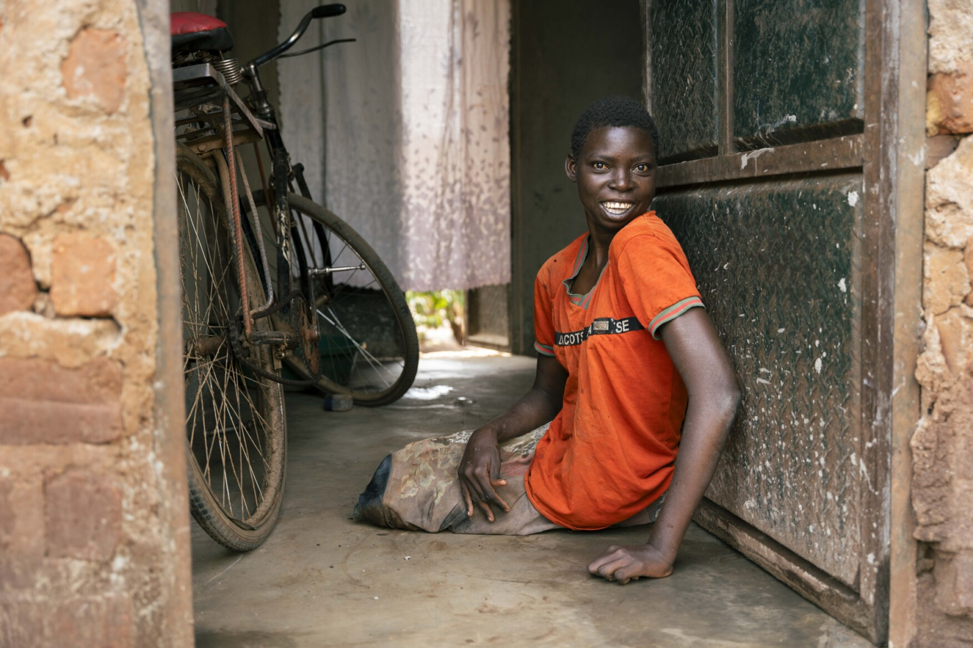 A woman sits on the floor in a doorway beside a bicycle inside a brick-walled passage, smiling at the camera.