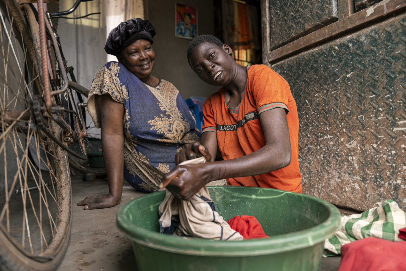 Two people sit by a doorway while washing clothes in a green tub; a bicycle frame is visible on the left.