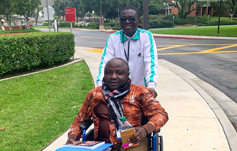 Daniel Kyungu in a wheelchair with a man pushing him.