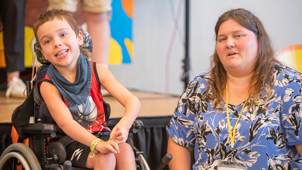 A smiling boy in a wheelchair next to a woman in a blue floral shirt at a stage event.