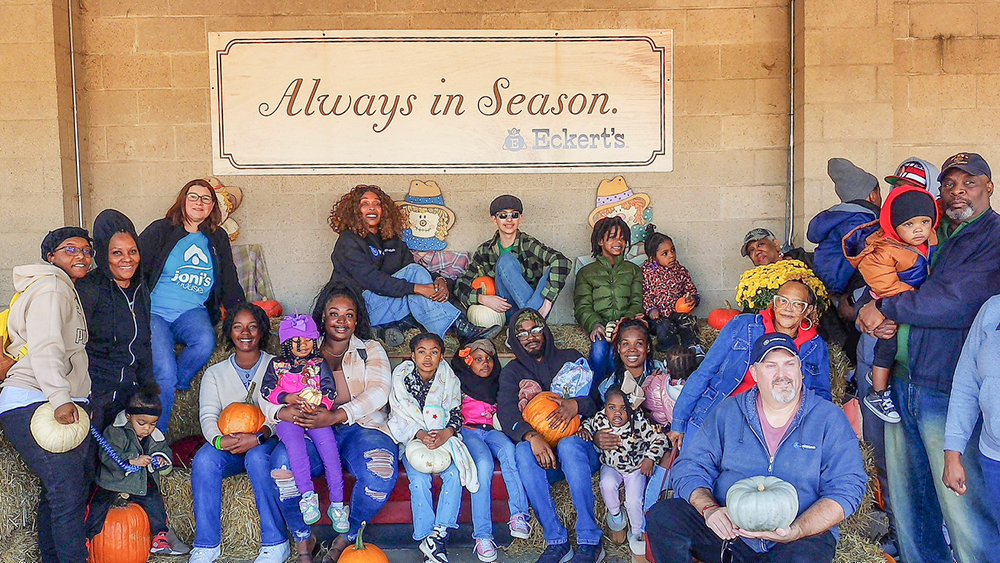 Group of diverse adults and children posing with pumpkins on hay bales under a sign reading 'Always in Season'.