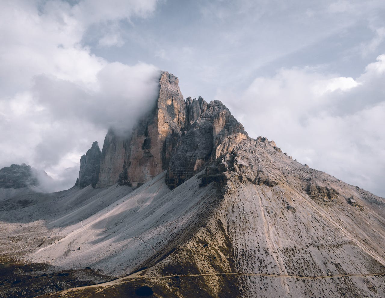 Jagged mountain peak rising from light grey slopes, with low clouds wrapping the summit and a dusty road at the base.
