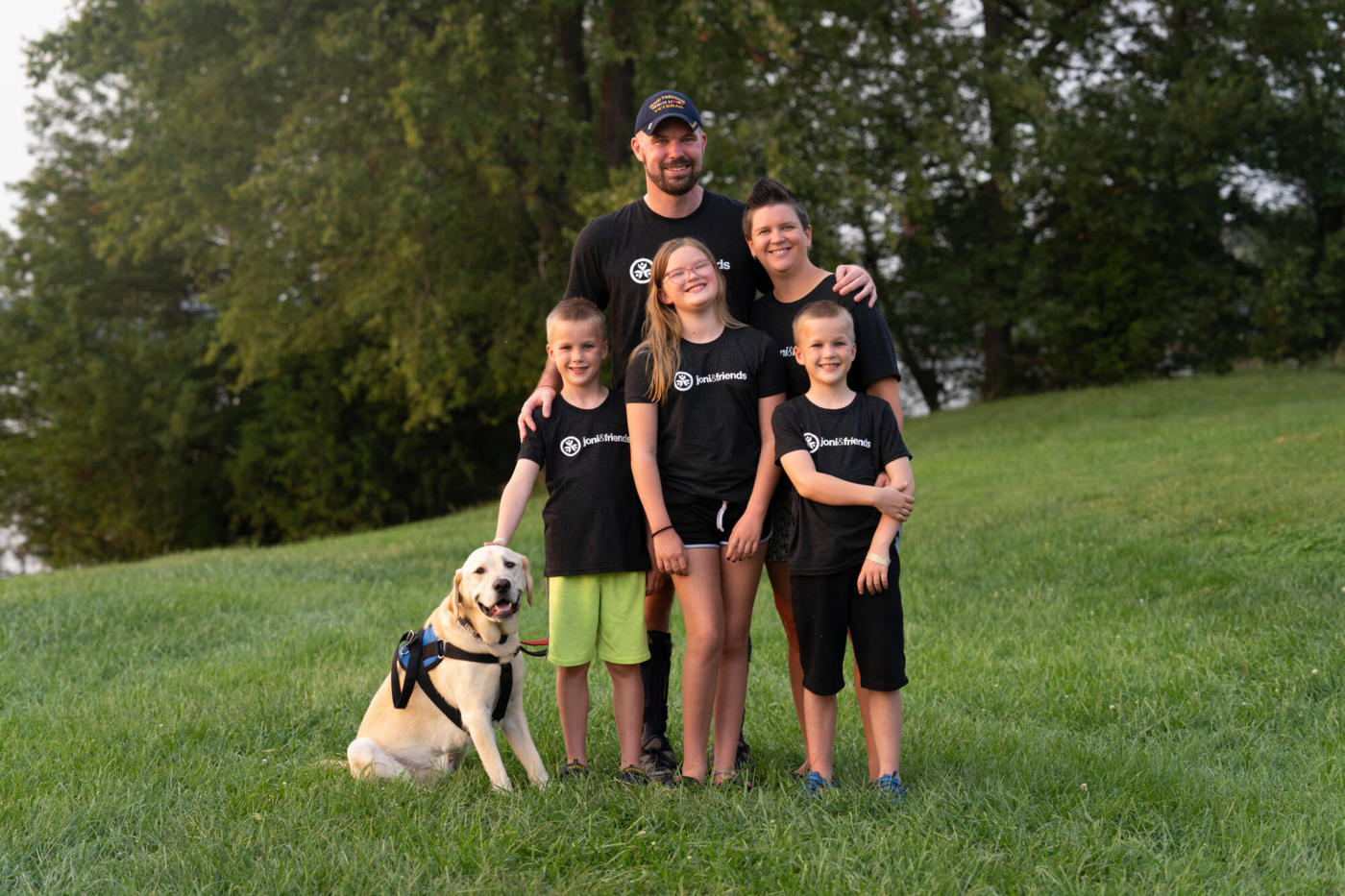 Matt and Kristi with their three children and a dog are posing and smiling for the camera outdoors on grass, with trees in the background.