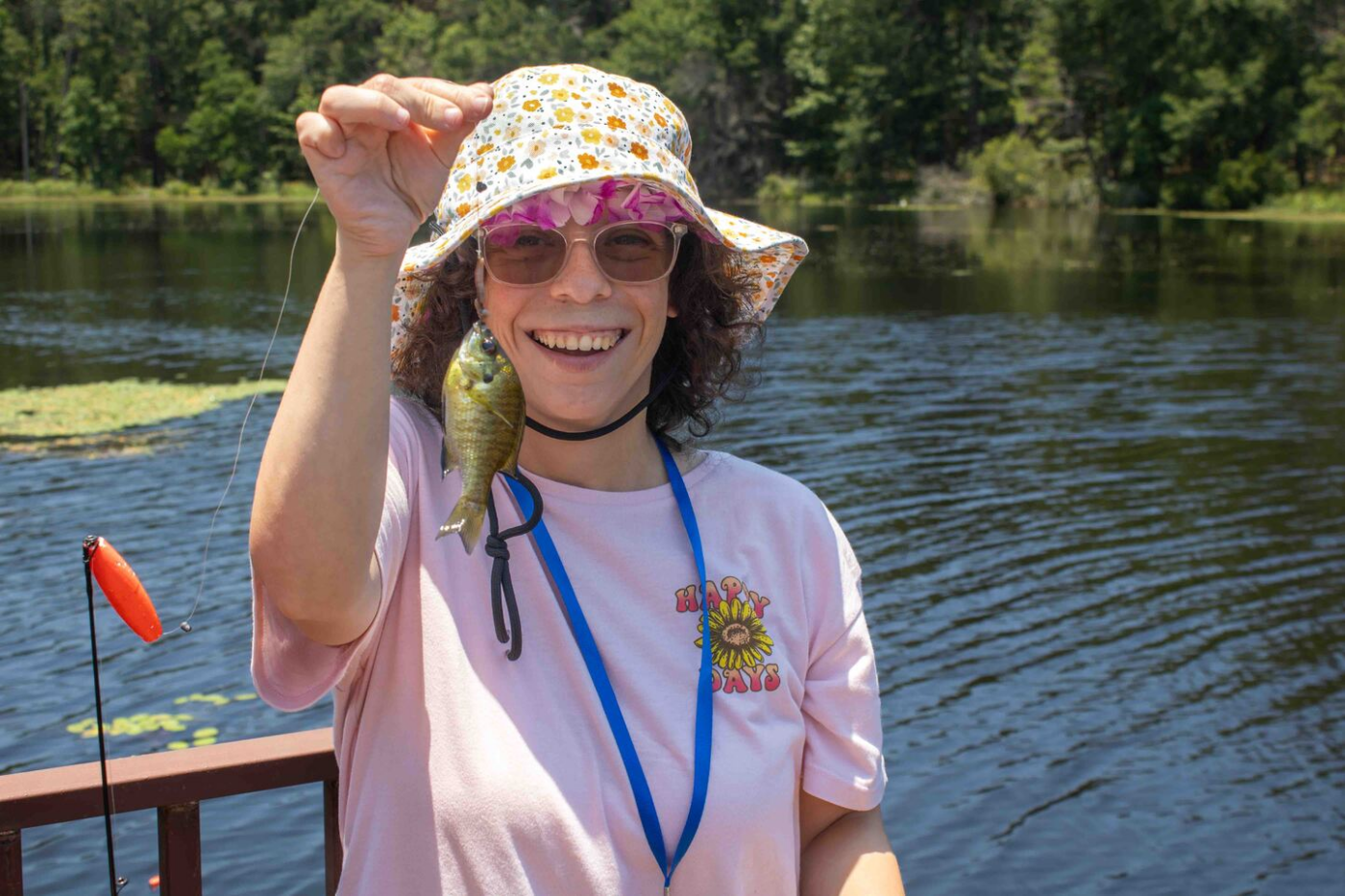 Bailey smiles while holding up a small fish by a lake, wearing a floral bucket hat, sunglasses, and a pink shirt with a sunflower logo.