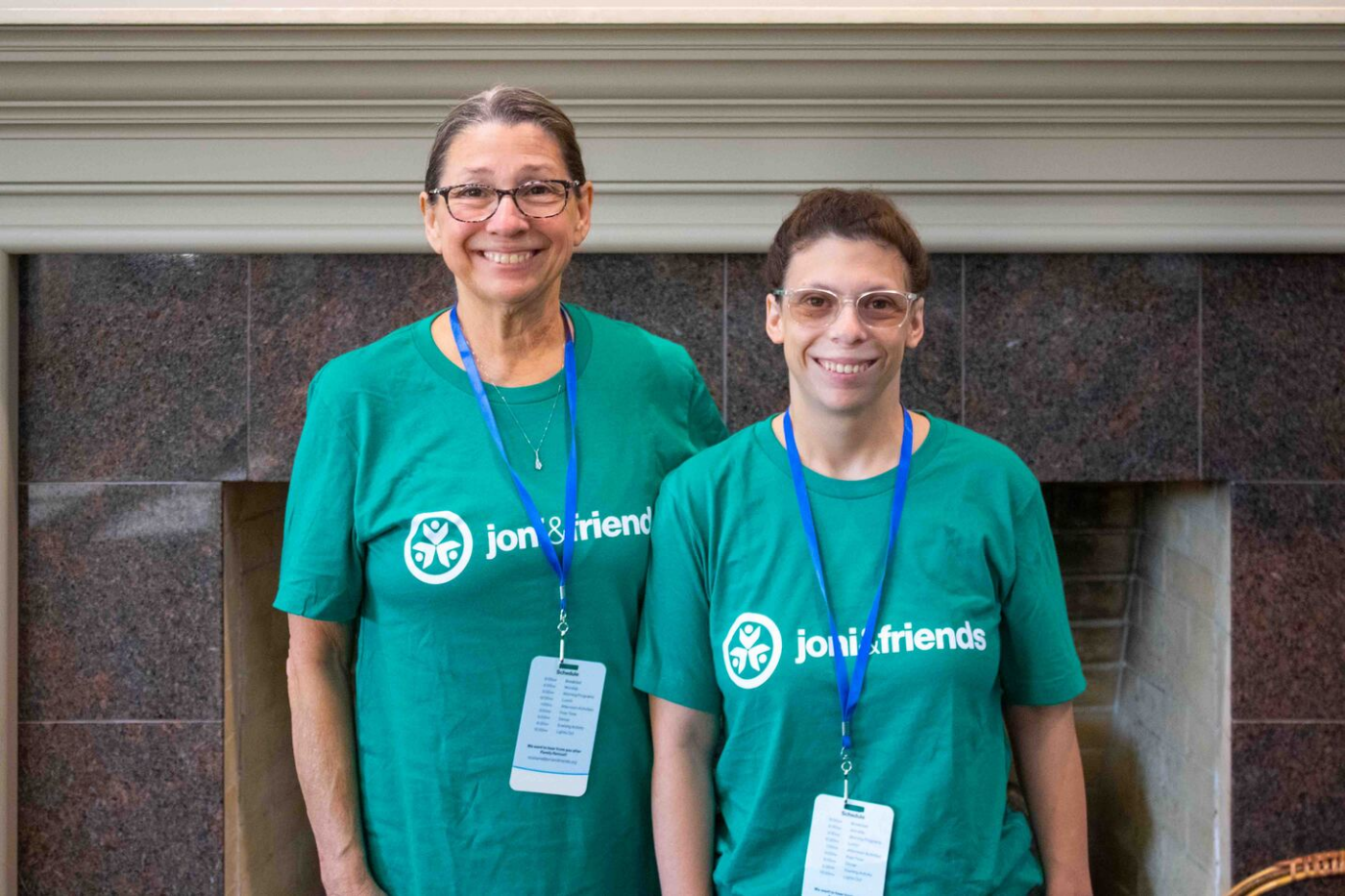 Lorna and Bailey wear Joni & Friends T-shirts with blue lanyards and name badges, standing next to each other in front of a fireplace.