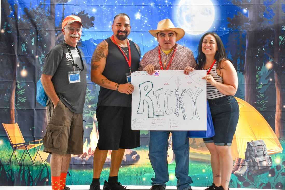 Ricky poses indoors with three other adults, holding a large sign that reads "RICKY," with a camping backdrop and a moonlit scene behind them.