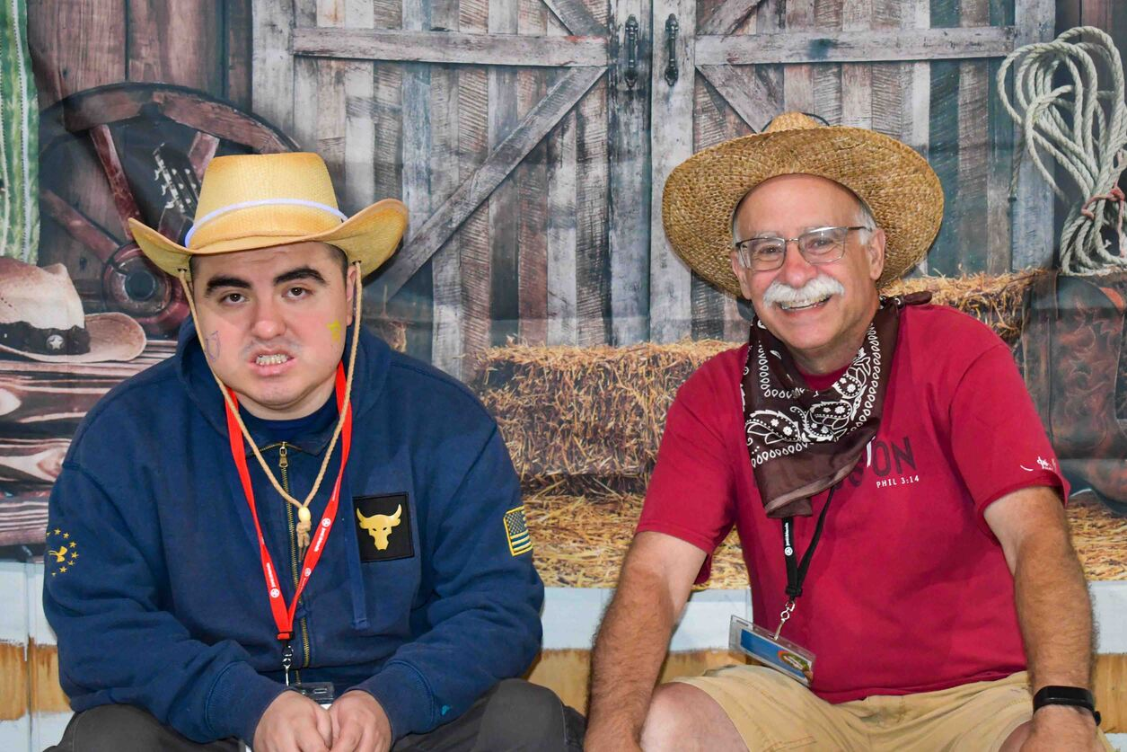 Ricky, wearing a blue jacket with a bull patch, a red lanyard, and a cowboy hat, smiles while seated beside another man in front of a rustic barn backdrop.