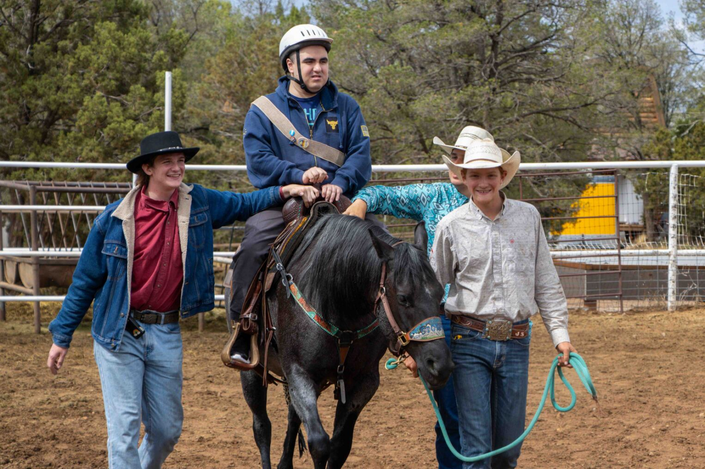 Ricky rides a dark horse, wearing a helmet and smiling, while three other people walk beside the horse and help guide it.