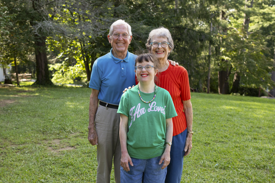 Ron, Cherry, and Jenny smile and pose for a family photo on grass with tall trees in the background.