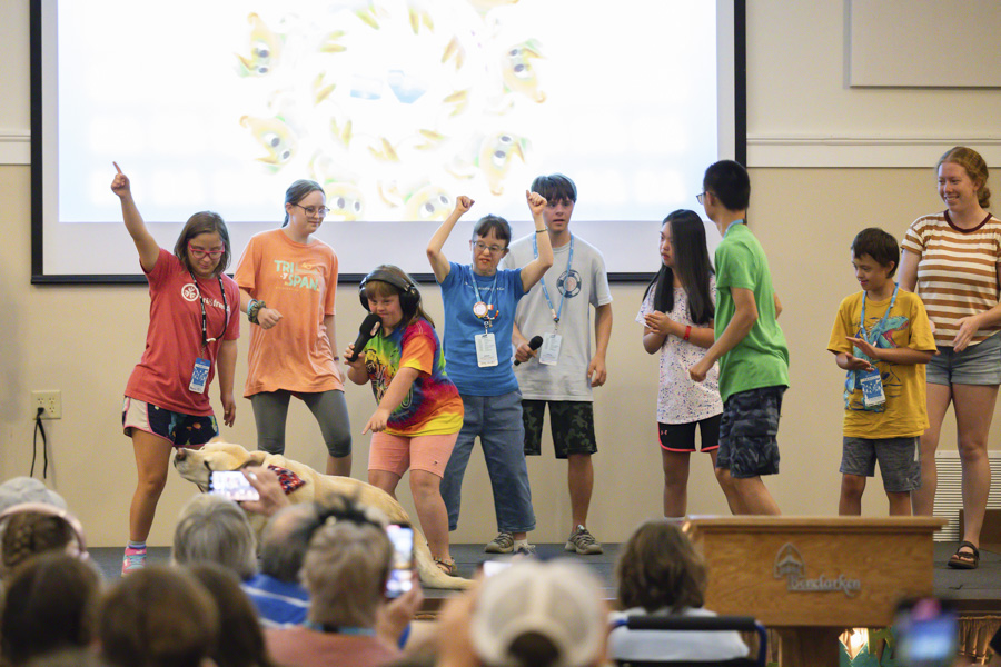 Jenny dances on a stage with other children during a performance featuring a dog, with an audience watching in front.