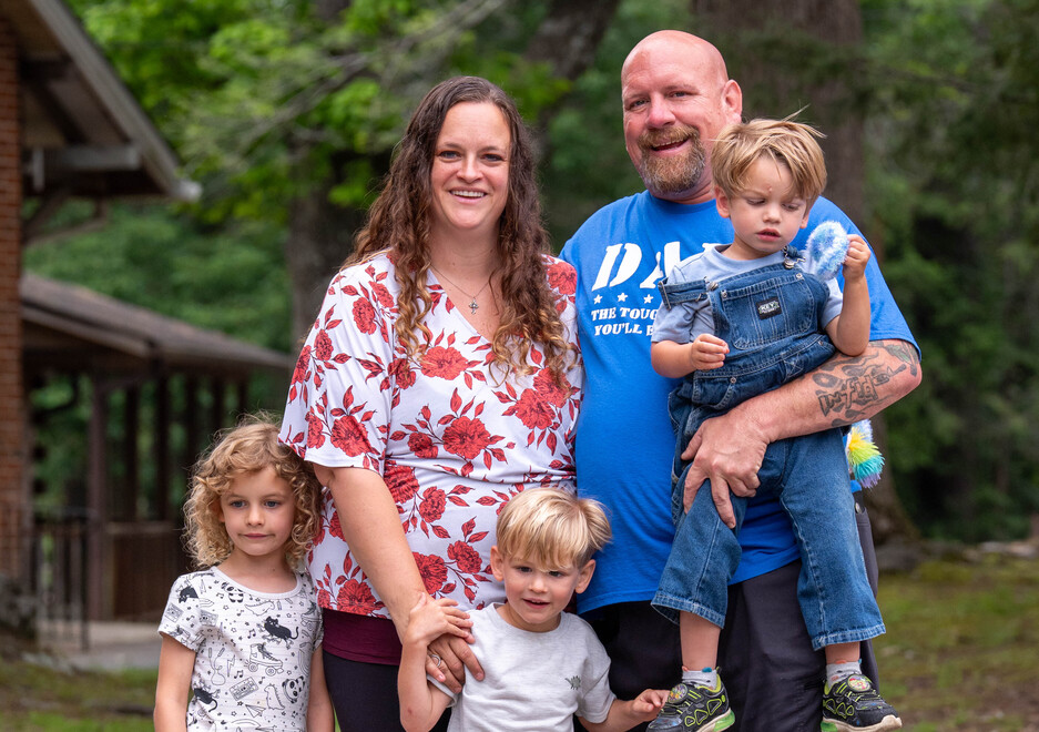 Julia and Andy Butterworth are smiling and posing outdoors with their two sons and daughter, with trees and grass in the background.