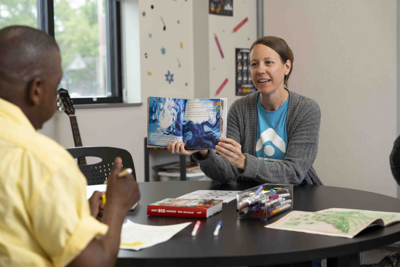 Woman shows a colorful picture book to a man at a round table indoors.