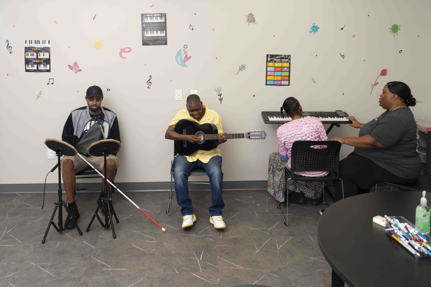 Four people in a music room: a man playing percussion, a man playing guitar, a woman at a keyboard, and another woman supervising.