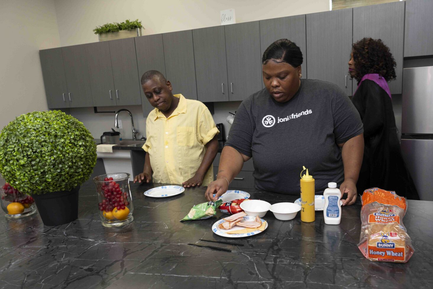A woman prepares a sandwich in a kitchen while a man stands beside her and another woman is in the background.
