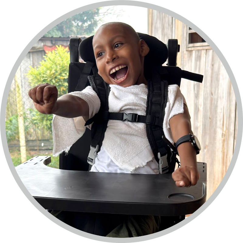 A child smiles while sitting in his new wheelchair on a wooden porch, wearing a white shirt and green pants, with a black table attached to the wheelchair.