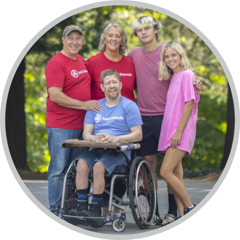 Five people pose outdoors for the camera, smiling. A man using a wheelchair is at the center of the group.
