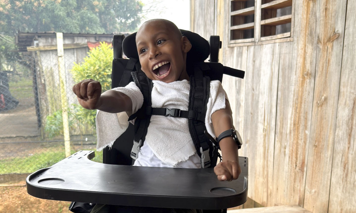 A child smiles while sitting in his new wheelchair on a wooden porch, wearing a white shirt and green pants, with a black table attached to the wheelchair.
