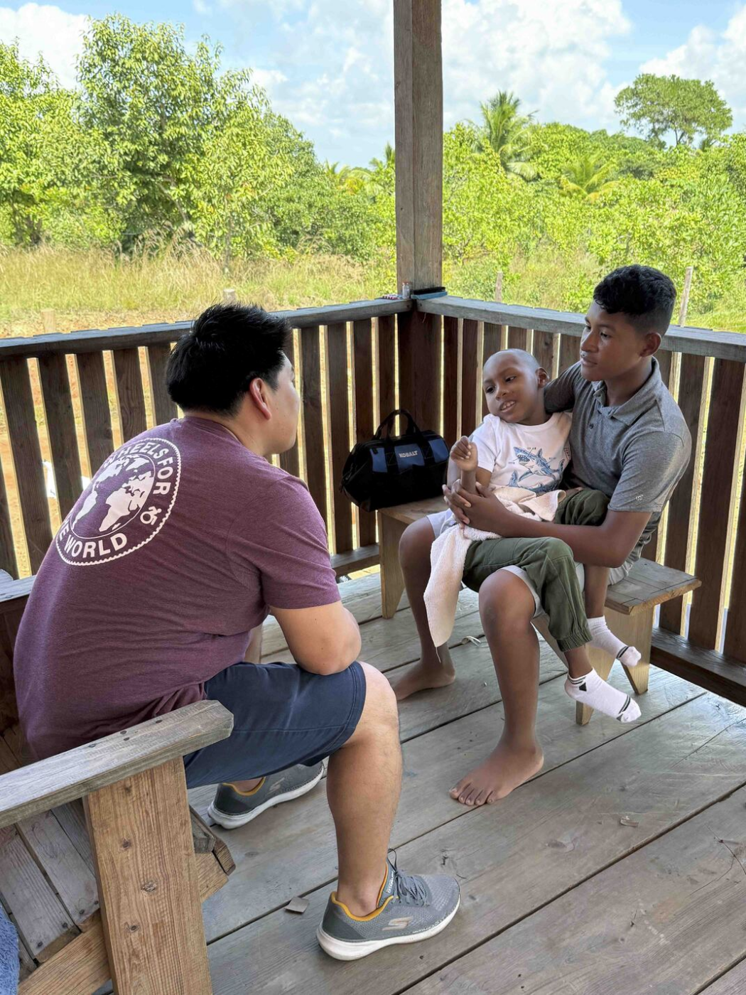 A man sits on a wooden porch surrounded by trees while talking to another man holding a child.