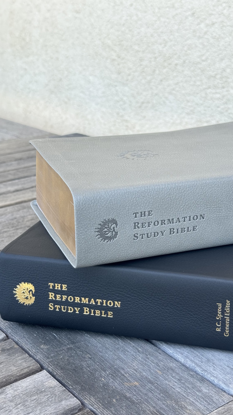 Two study Bibles stacked on a wooden surface: a gray Reformation Study Bible on top of a black one, both with lettering and embossed logos.