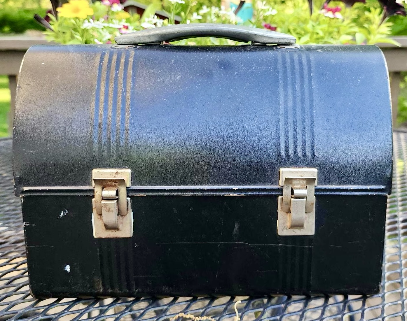 Black metal toolbox with two metal latches and a worn handle sits on a metal outdoor table, with colorful flowers in the blurred background.