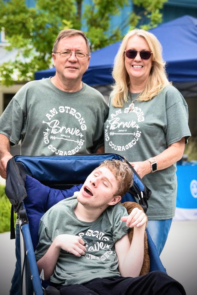 Ann and Bob are standing behind their son Joshua in an adult stroller. All are smiling for the camera.