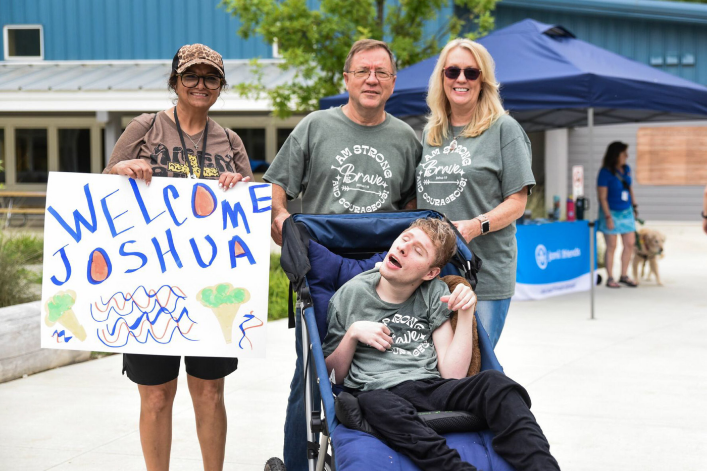 A woman holds a white cardboard sign that says "Welcome Joshua" beside Ann and Bob, who are standing behind their son Joshua in an adult stroller. All are smiling for the camera.