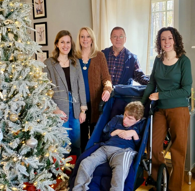 Joshua sits in an adult stroller with Ann and Bob and two other women in a group photo inside a home, with a Christmas tree in the background.