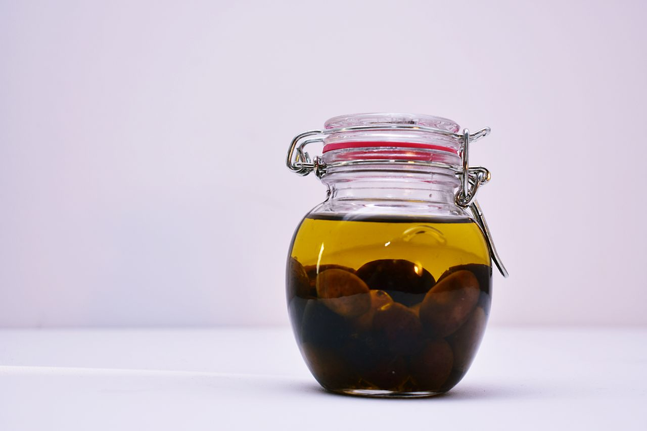 Glass jar of olive oil with whole olives settled at the bottom, sealed with a metal clasp on a white surface.