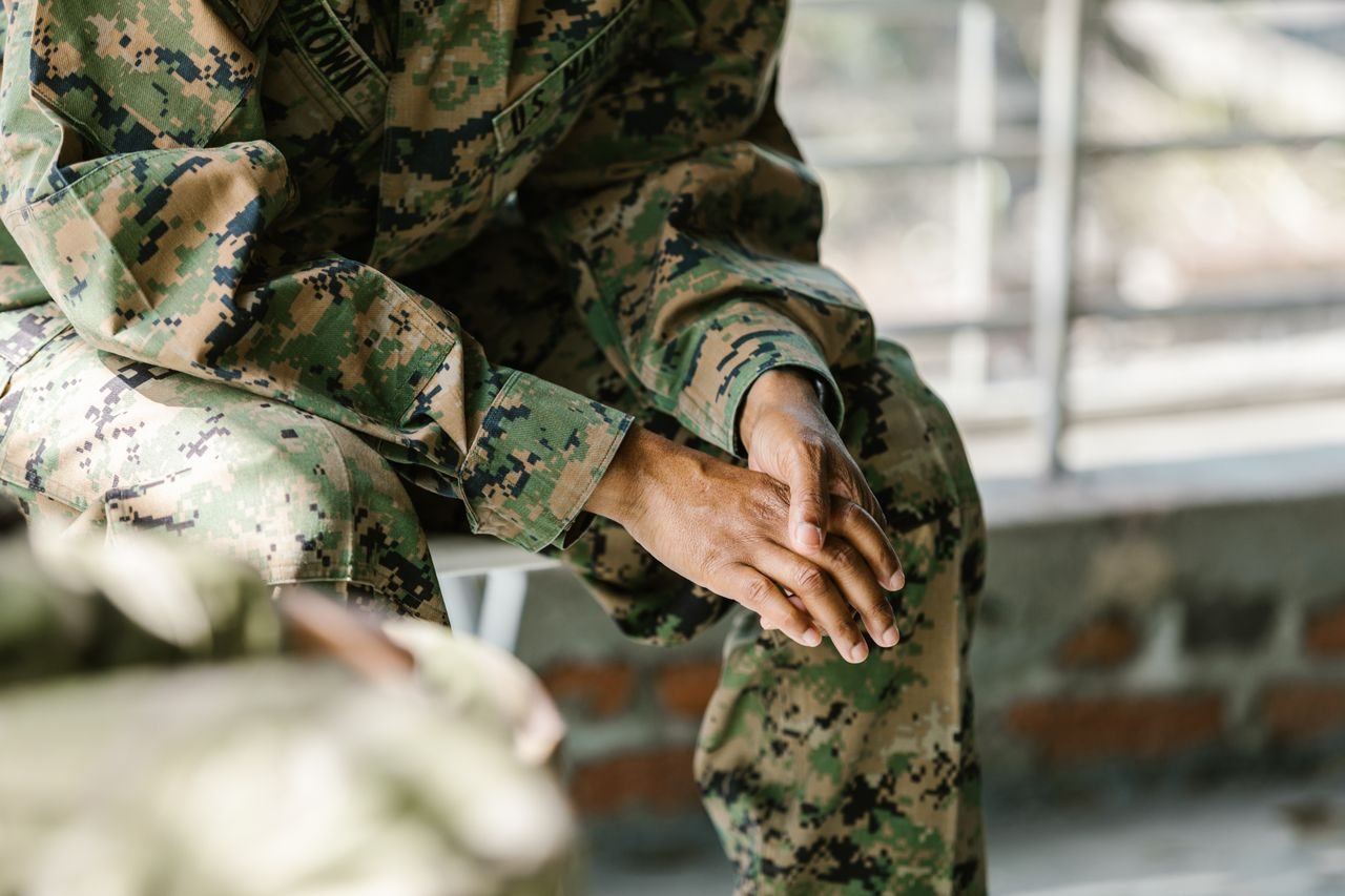 Soldier in camouflage uniform sitting outdoors with hands clasped in lap, resting.