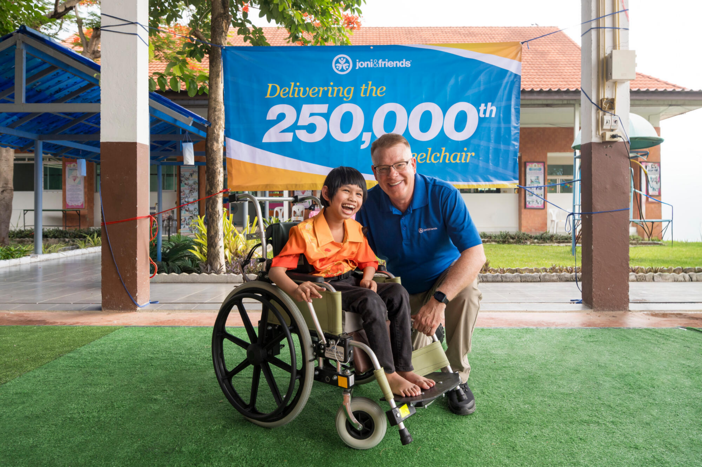 Shawn Thornton, president of Joni and Friends, next to Nada, a young girl with cerebral palsy, who is sitting in her new wheelchair. They both smile joyfully in front of a banner that reads “Joni & Friends Delivering the 250,000th Wheelchair.”