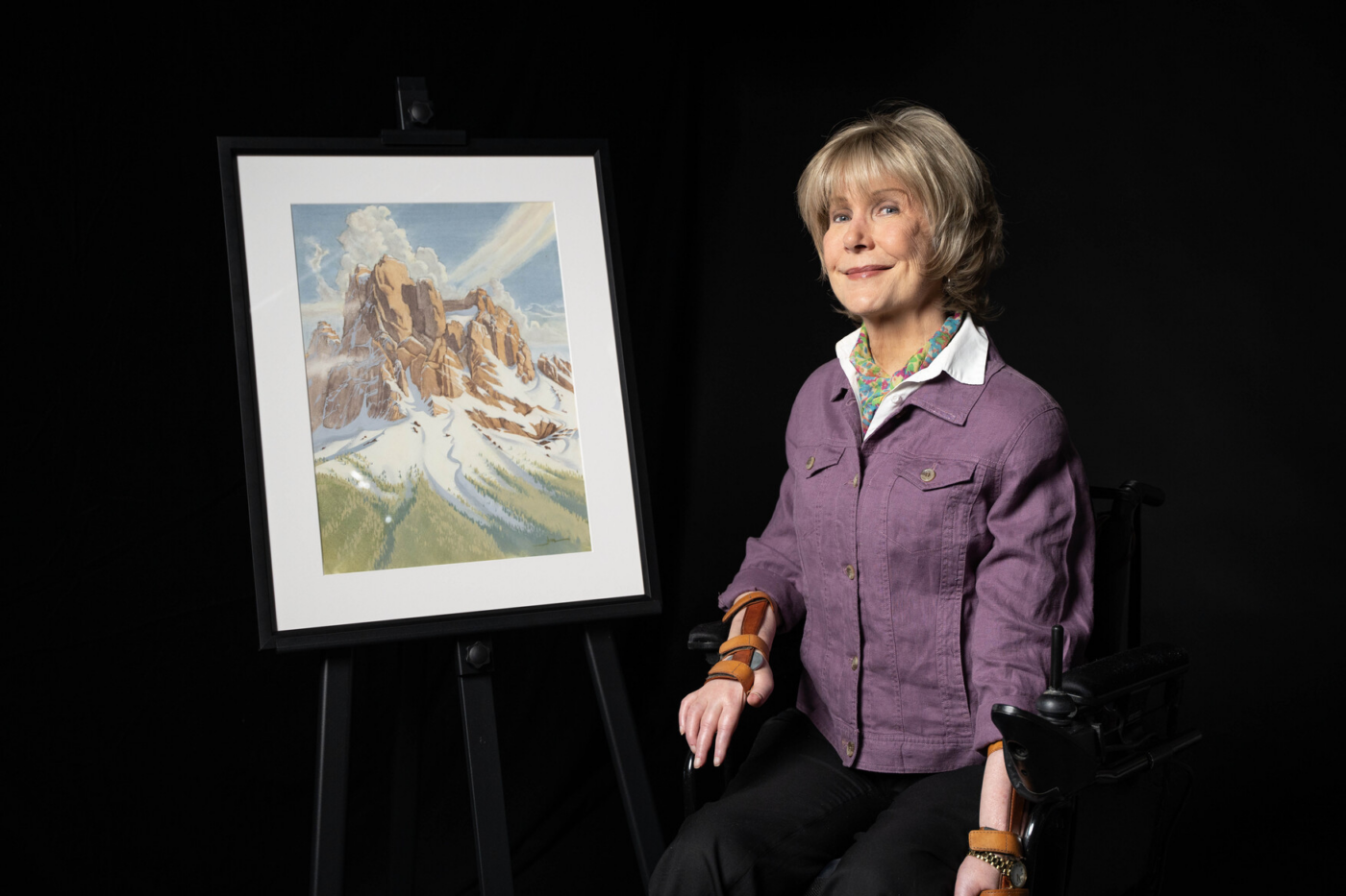 Joni Eareckson Tada beside her artwork “Rock of Ages,” showing towering sandstone boulders, snow, grass, and clouds.