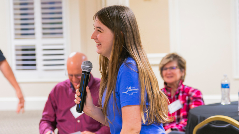 Woman in a blue shirt speaking into a microphone at a community event, with two seated panelists in the background