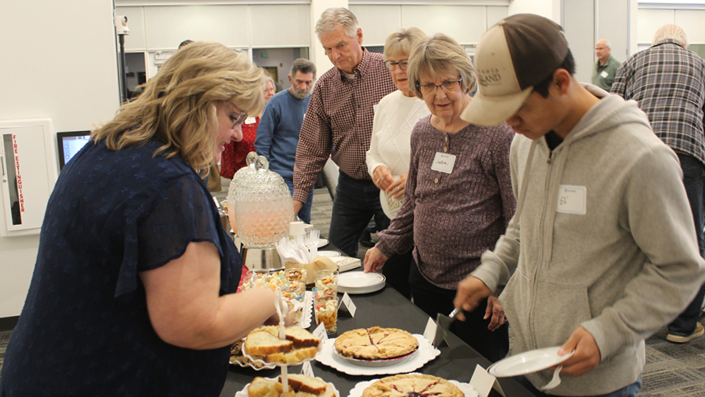 Group of adults gathered around a dessert table, selecting slices of pie at a community event.