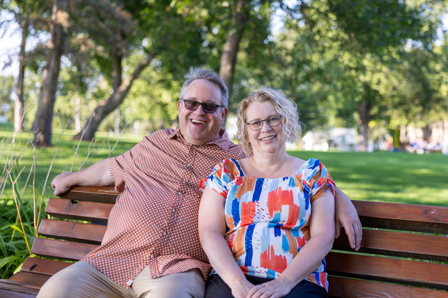 Hugh and Patricia sit on a wooden bench outdoors, smiling and posing for the camera.