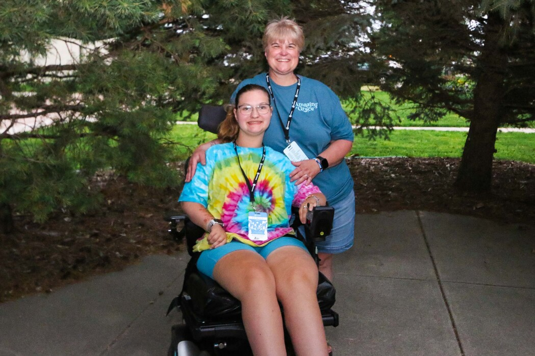 Abi in her wheelchair next to her mom, Erica. They are both smiling at the camera, with trees in the background.