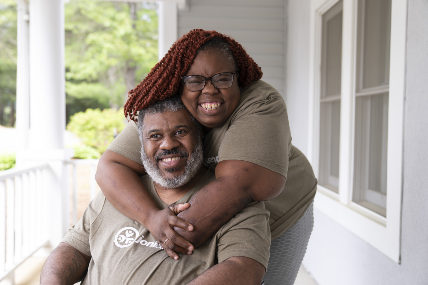 Smiling couple on a porch. Natacha hugs her husband from behind.